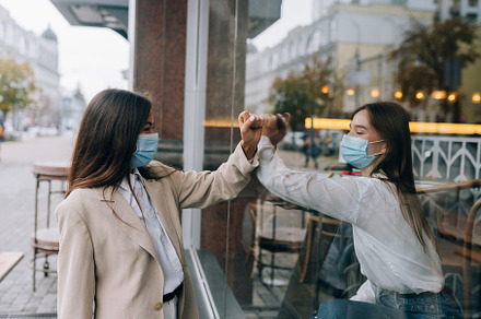 Two women in protective masks opposite each other, window between them.
