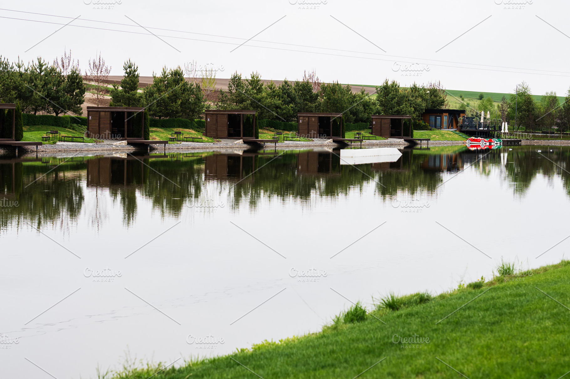 Row of outdoor huts reflected on a calm lake surface on a cloudy day, a Photo by AS photostudio