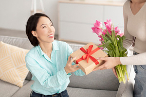 Young woman giving wrapped gift box and beautiful flowers to her mature Asian m, a Photo by Prostock-Studio