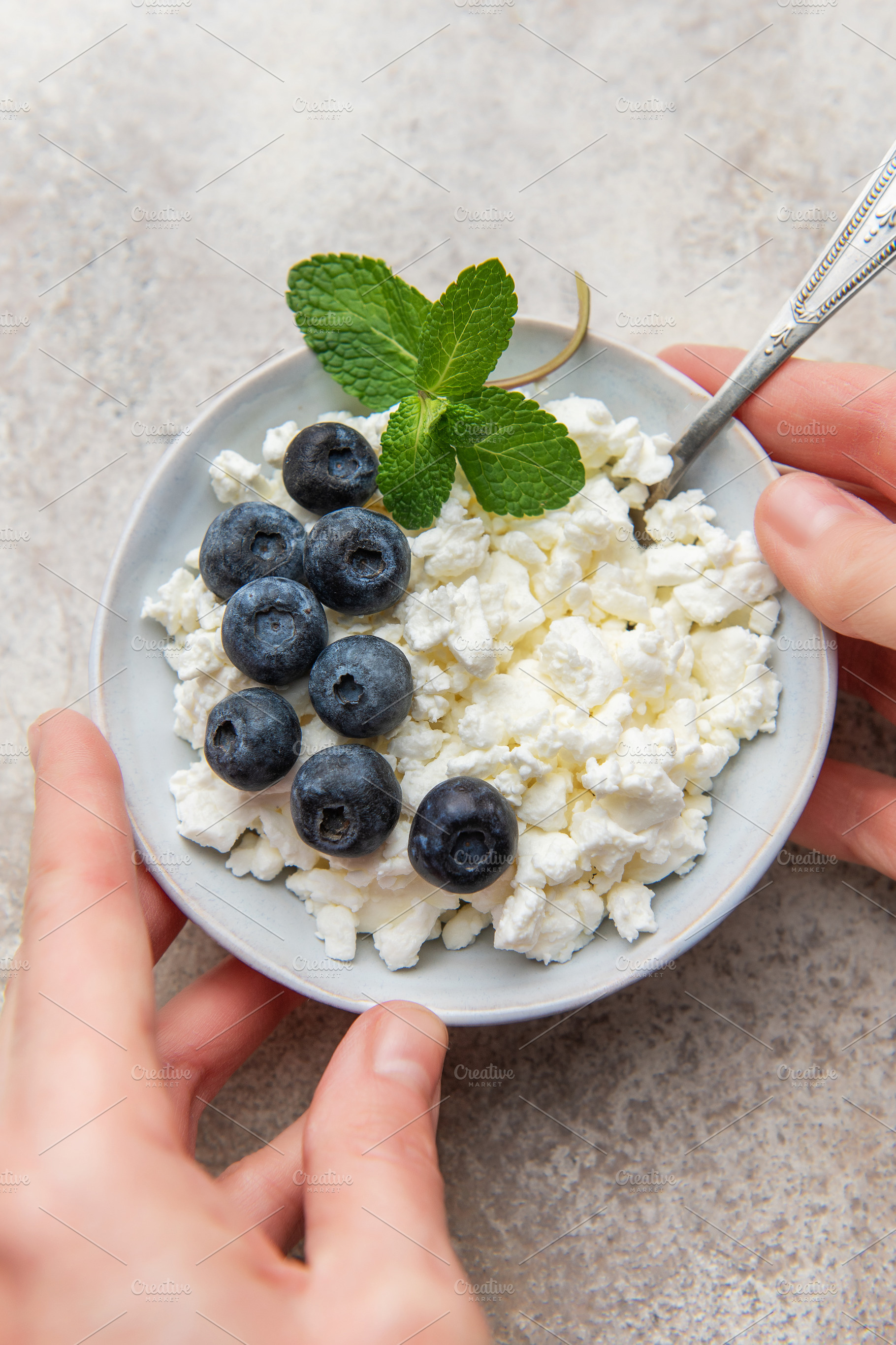 Hands holding a bowl of cottage cheese with blueberries and mint, a Food & Drink Photo by Olena Rudo