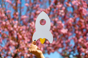 Hand holding a white rocket cutout with yellow flame in front of, a Nature Photo by Ales Munt