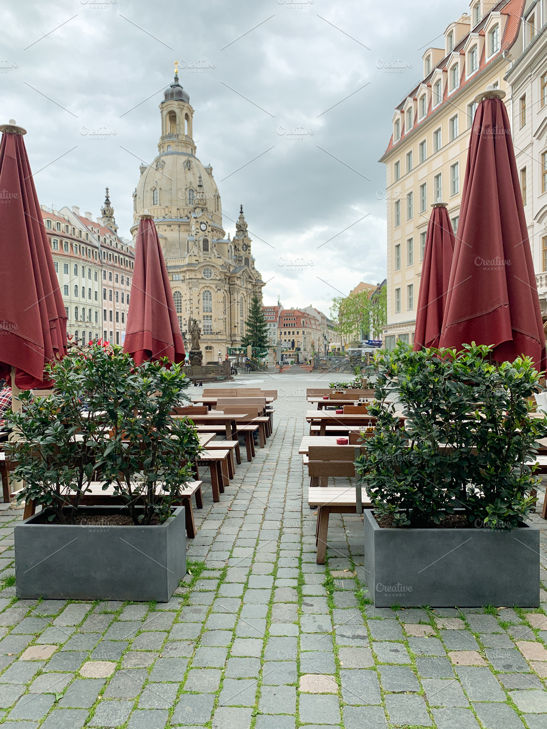 Outdoor café view with red umbrellas, benches, and green plants, a Holiday Photo by sebastianaraw