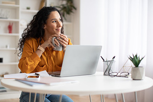 Dreamy Businesswoman Using Laptop And Holding Coffee Cup Indoors, a Photo by Prostock-Studio