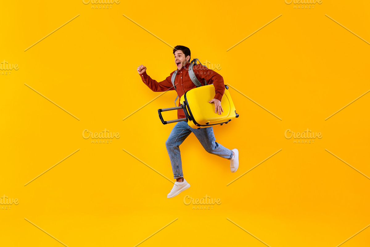 Emotional Male Tourist Jumping Carrying Suitcase Over Yellow Studio Background, a Photo by Prostock-Studio (Photo 2 of 2)