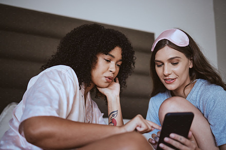 Phone, sleepover and friends in bed talking, bonding and networking on social media, mobile app or internet. Fun, technology and women browsing on a website with a cellphone in a bedroom at home., a Photo by Peopleimages.com