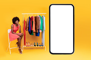 Black Woman Sitting Near Cellphone And Clothing Rack, Yellow Background, a Photo by Prostock-Studio