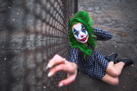 Greenhaired girl in chekered dress with joker makeup sitting near wire mesh fen