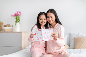 Happy asian teen girl and young woman reading postcard sitting on bed in minima, a Photo by Prostock-Studio