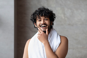 Portrait of happy bearded indian man with towel posing in bathroom and touching, a Person Photo by Prostock-Studio