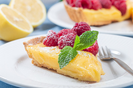 Piece of lemon tart on white plate, garnished with fresh raspberry, icing sugar, a Food & Drink Photo by iuliia_n