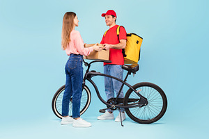 Courier Delivering Parcel To Woman Posing With Bike, Blue Background, a Photo by Prostock-Studio