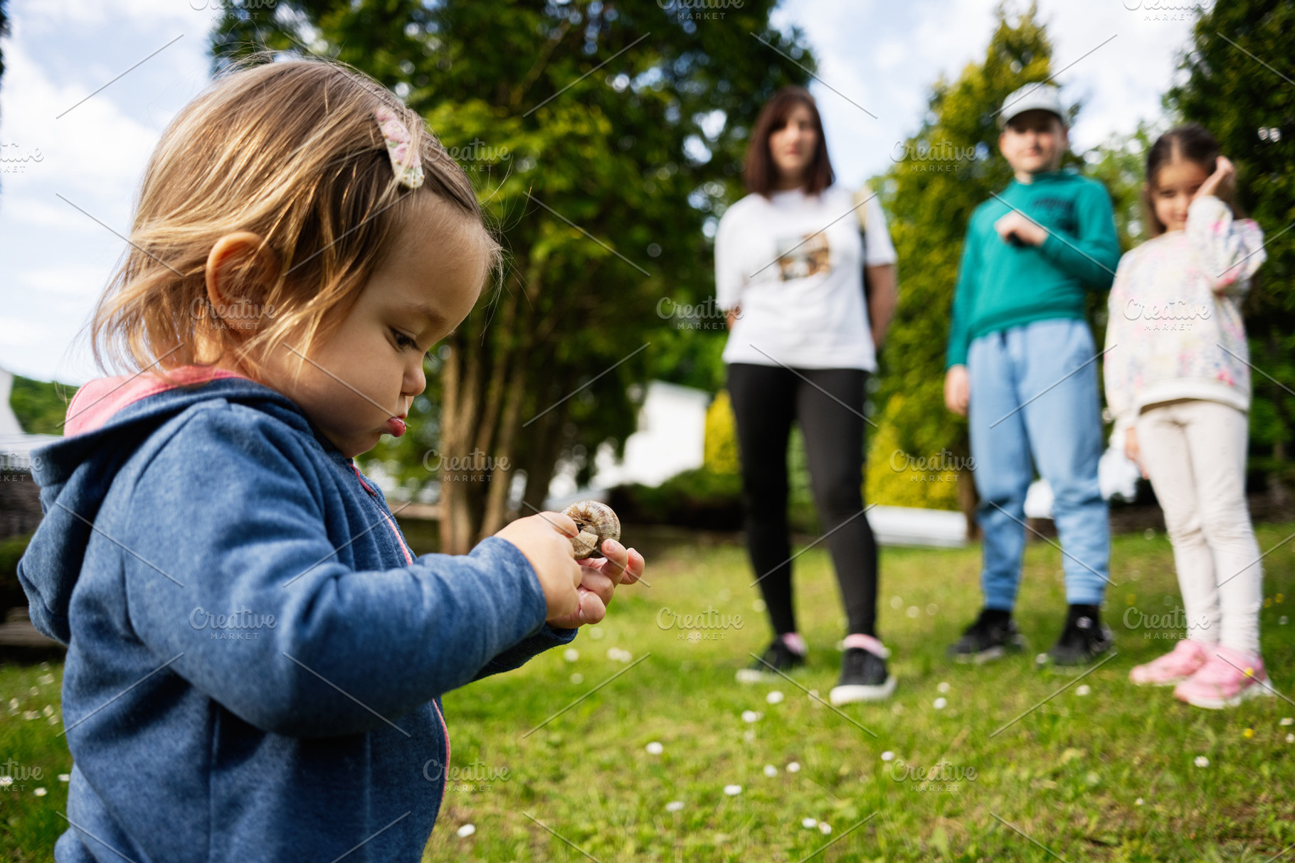 Young child holding an object outdoors with family in blurred background, a Photo by AS photostudio