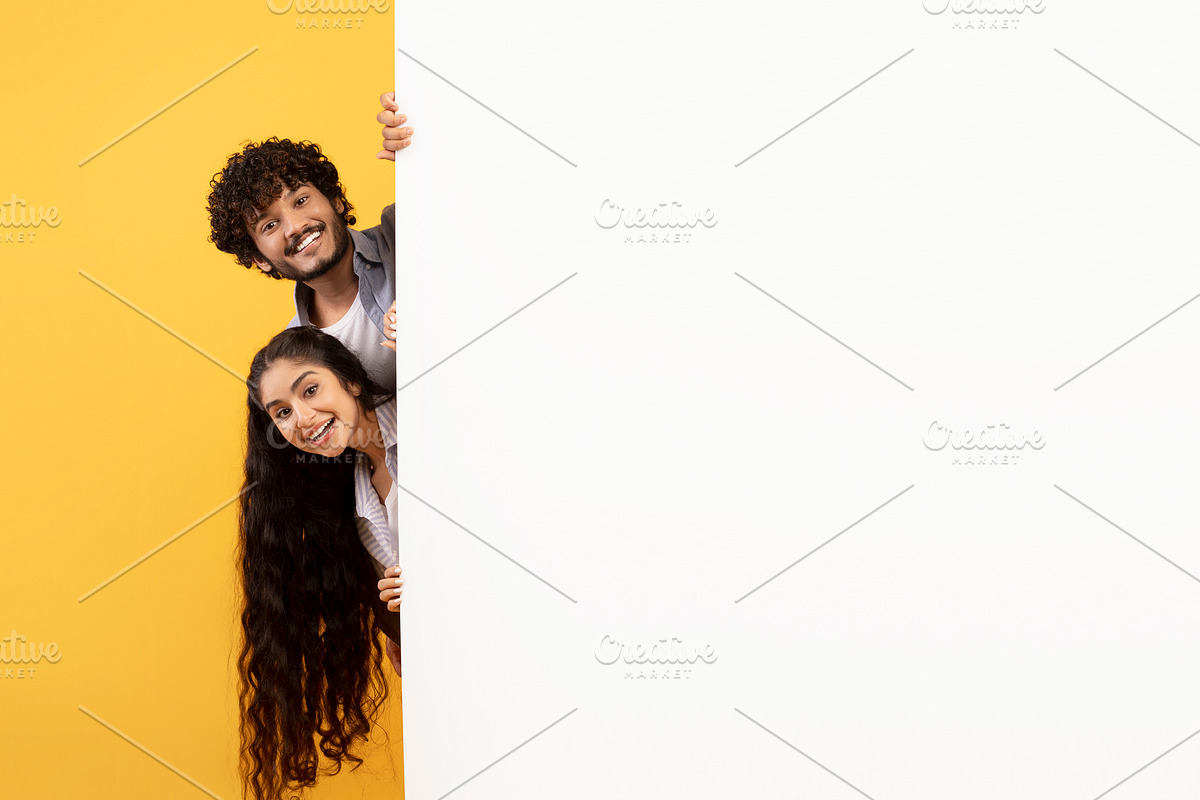 Excited indian couple standing behind white advertisement board and peeking out, a Photo by Prostock-Studio (Photo 2 of 2)