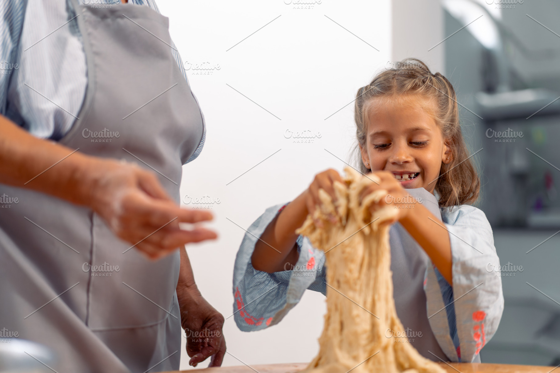 Happy girl kneading dough while baking with grandmother at home, a Photo by andreonegin market
