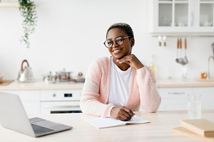 Happy young black lady in glasses and casual sits at table looks at laptop make, a Person Photo by Prostock-Studio