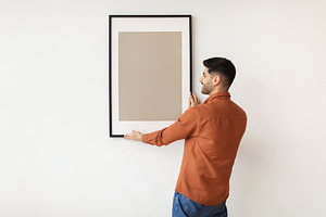 Young man hanging picture frame on the wall, a Photo by Prostock-Studio