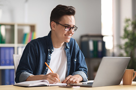 Happy millennial businessman taking notes while working with laptop in office