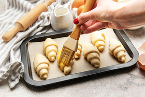 Baker brushing egg yolk on raw croissants on baking sheet, a Food & Drink Photo by Olena Rudo