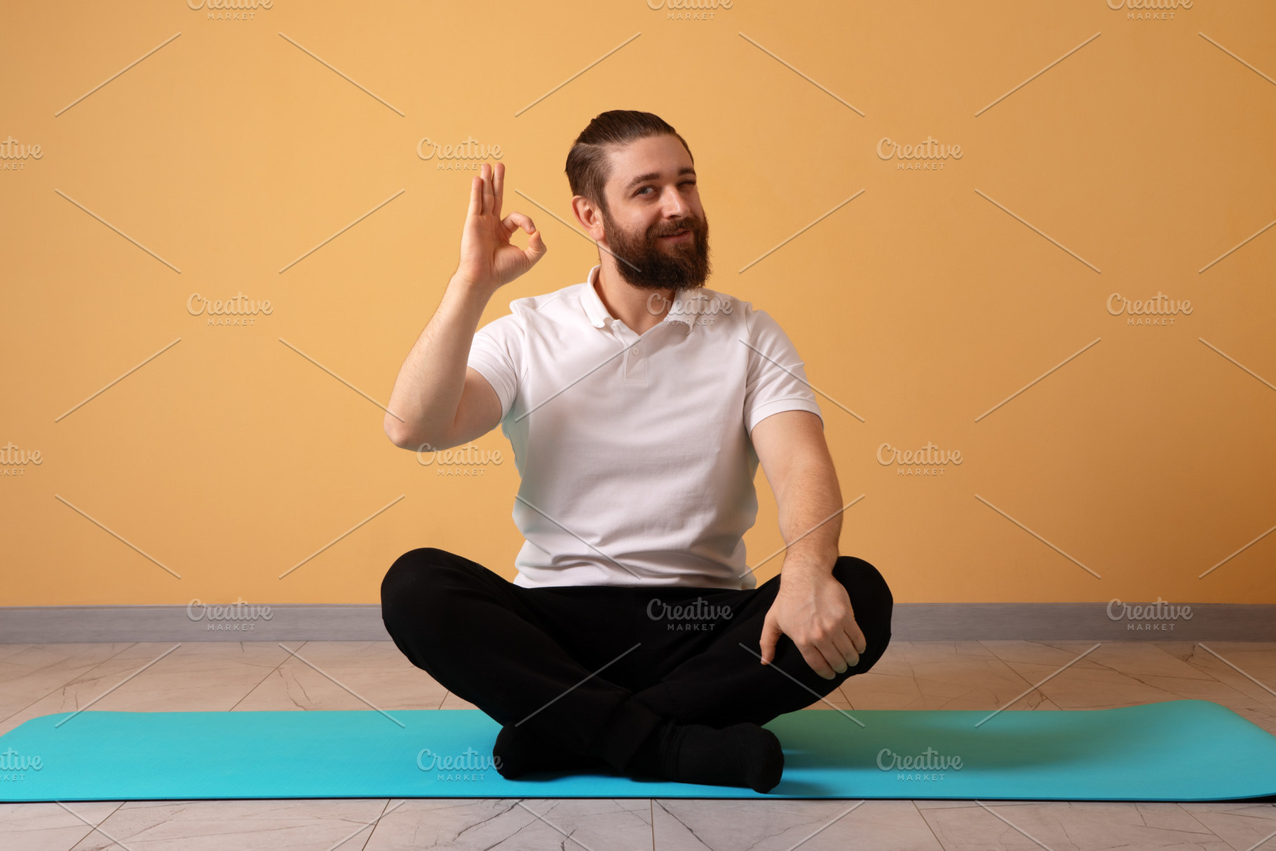 Bearded man sitting cross-legged on a yoga mat, making an OK ges, a Person Photo by Ales Munt