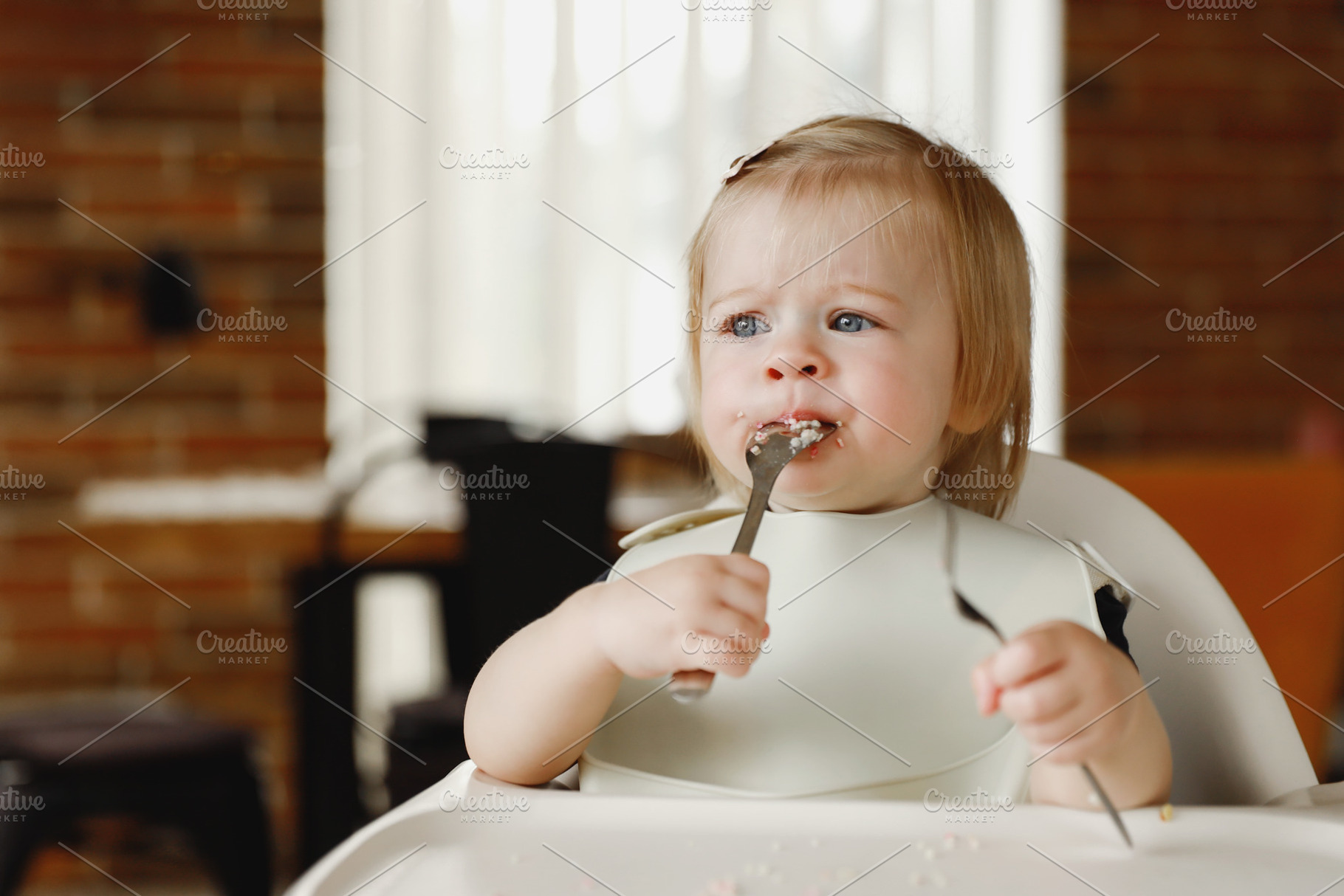 Infant girl or toddler sitting on high chair. Eating food by herself using spoo, a Person Photo by Lenor4ik