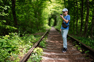 Boy walking on railway tracks and using mobile phone in green forest, a Photo by AS photostudio