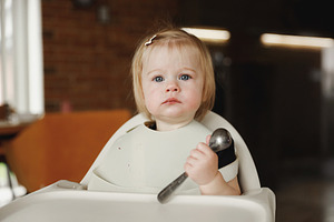 Infant girl or toddler sitting on high chair. Eating food by herself using spoo