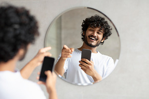 Excited indian guy taking photo in bathroom mirror holding smartphone and point, a Person Photo by Prostock-Studio