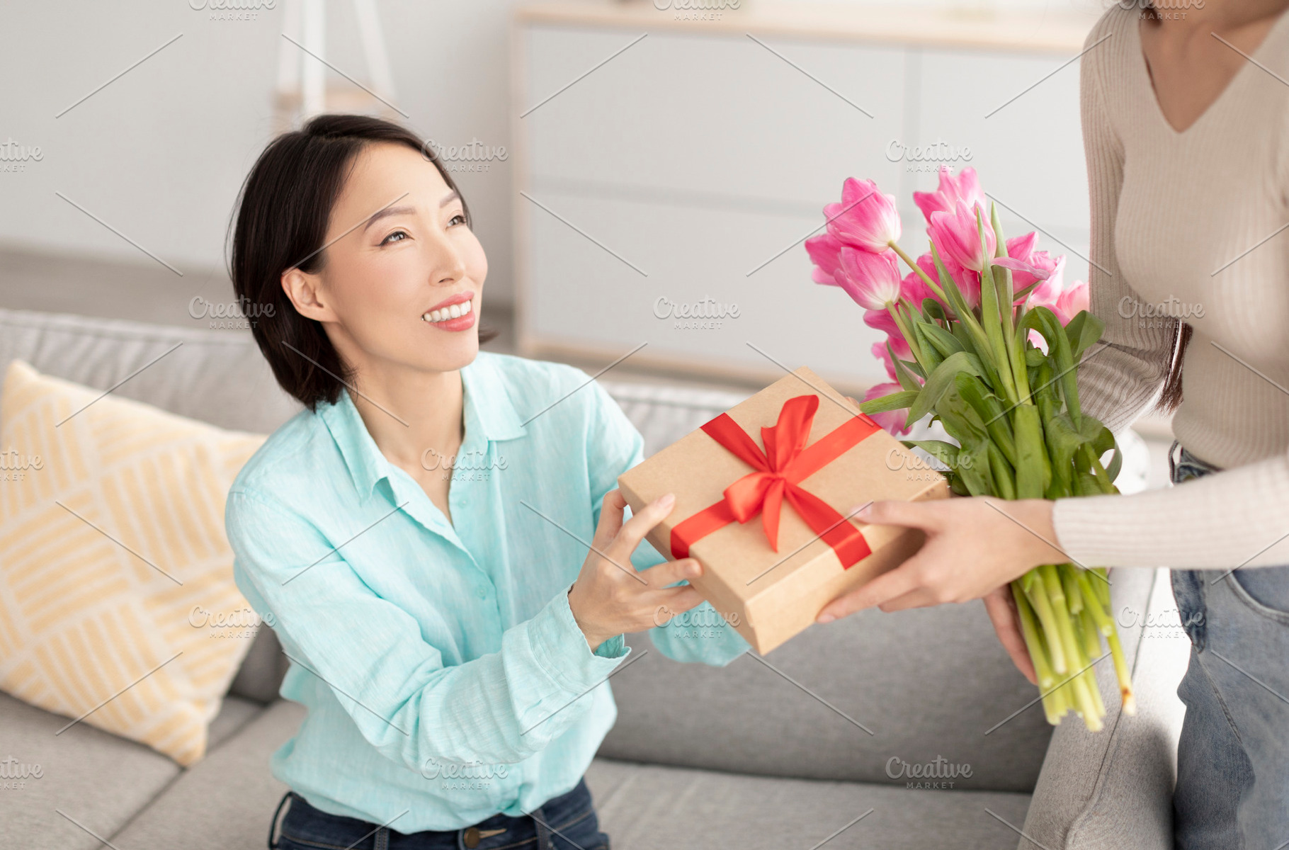 Young woman giving wrapped gift box and beautiful flowers to her mature Asian m, a Photo by Prostock-Studio