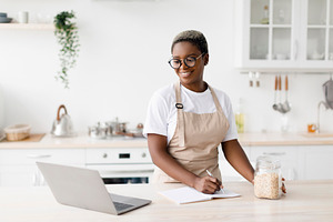 Cheerful millennial pretty black female in glasses, apron writes recipe looks p, a Person Photo by Prostock-Studio