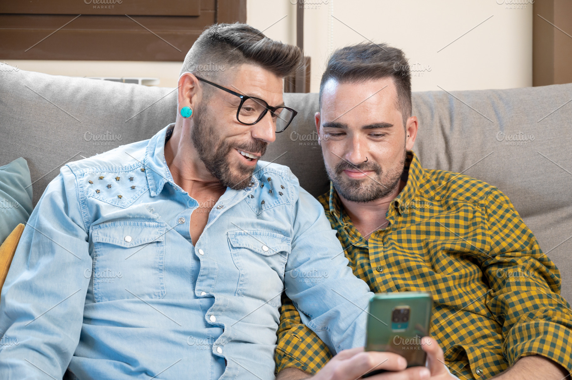 Happy young gay couple using mobile phone while sitting on a sofa in the living, a Person Photo by david.herraez.calzad