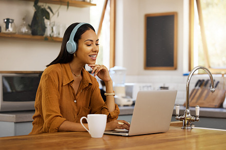 Young happy mixed race businesswoman talking while on a virtual meeting using a laptop at home. One hispanic female businessperson talking and wearing headphones while on a call using a laptop, a Photo by Peopleimages.com