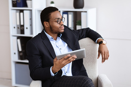 Thoughtful black businessman sitting in chair, using digital tablet, a Photo by Prostock-Studio