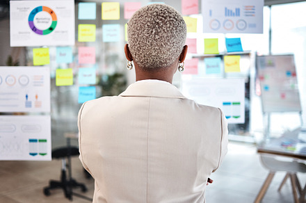Business woman, back and thinking in project management, marketing or planning at the office. Rear view of Female person, manager or employee checking vision board of company statistics at workplace, a Photo by Peopleimages.com
