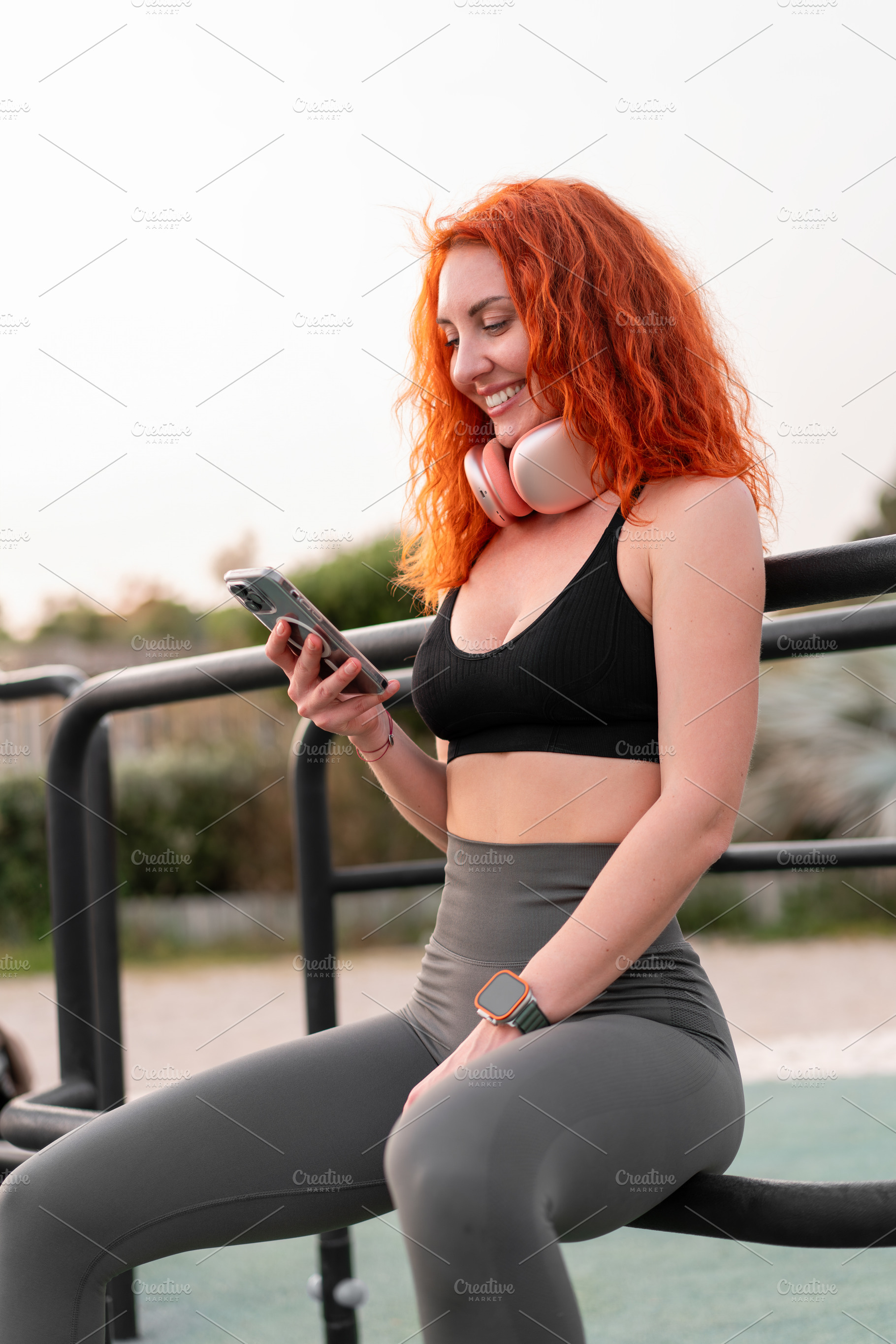 Young woman with red hair checks phone while exercising outdoors in fitness gea, a Person Photo by andreonegin market