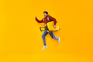 Emotional Male Tourist Jumping Carrying Suitcase Over Yellow Studio Background, a Photo by Prostock-Studio