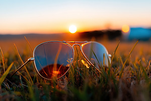 Sunglasses on grass during sunset with warm hues illuminating the evening sky, a Beauty & Fashion Photo by AlyaAnd