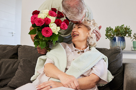 Couple kiss, senior and flowers for a birthday, love or celebration of marriage together. Happy, care and elderly man with bouquet for a woman in a house for a surprise, date or gift for anniversary, a Photo by Peopleimages.com
