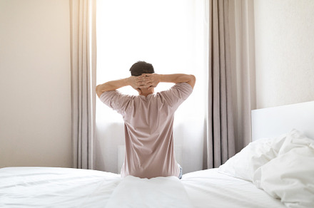 Back view of fully-rested man sitting on bed, stretching, a Photo by Prostock-Studio