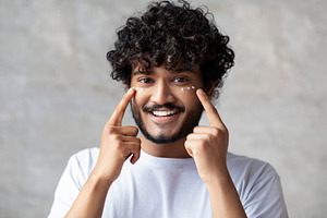 Portrait of happy indian man applying cream under eyes and smiling at camera, e, a Person Photo by Prostock-Studio