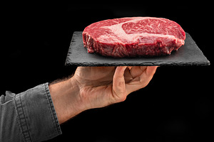 Cook holds graphite serving board with raw ribeye steak on black, a Food & Drink Photo by Volff