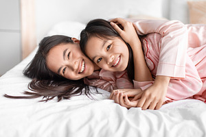 Glad happy pretty cute japanese young woman hug teen girl in pink pajama lie on, a Photo by Prostock-Studio