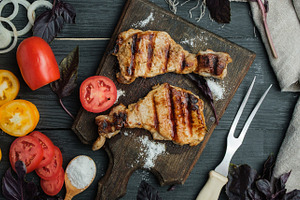 Grilled pork steaks on a wooden board, surrounded by vegetables, a Food & Drink Photo by ndanko