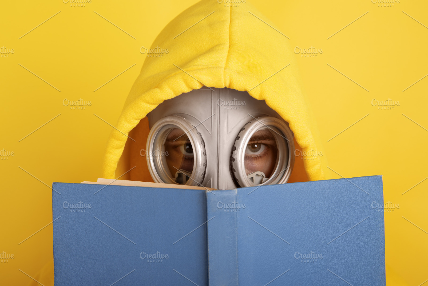 Person in a yellow hoodie and gas mask reading a blue book again, a Person Photo by Ales Munt