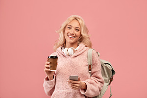 Pretty lady posing with backpack, holding coffee to go and cellphone, smiling a, a Photo by Prostock-Studio
