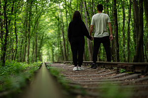 Couple holding hands walking along a forest path between trees on train tracks, a Photo by AS photostudio