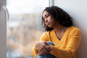 Depressed young African American woman sitting near window, looking outside on, a Photo by Prostock-Studio