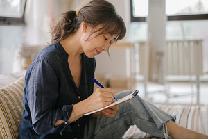 Asian woman making notes in notebook, working at home on bed in cozy bedroom. C