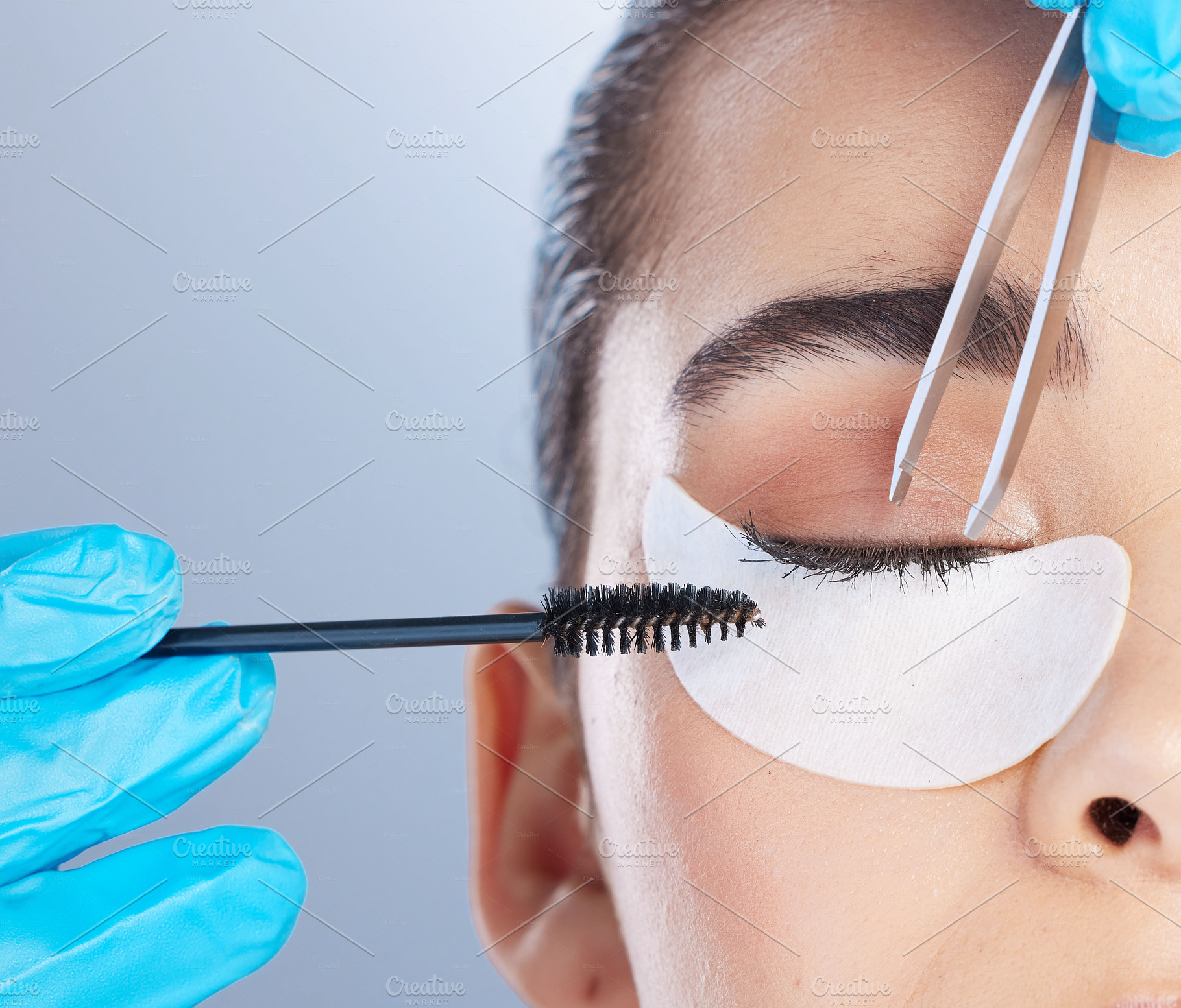 Woman, lashes and tweezer with mascara, studio closeup and makeup artist in gloves, hands and extension. Girl, model and zoom of face for beauty, cosmetics and brush for aesthetic by gray background, a Photo by Peopleimages.com