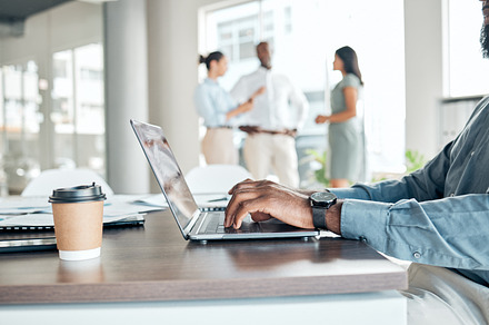 Laptop, coffee and a business man at a startup working at a desk in an open office with documents. Teamwork, planning and internet research, a manager at a company with a team talking in background., a Photo by Peopleimages.com