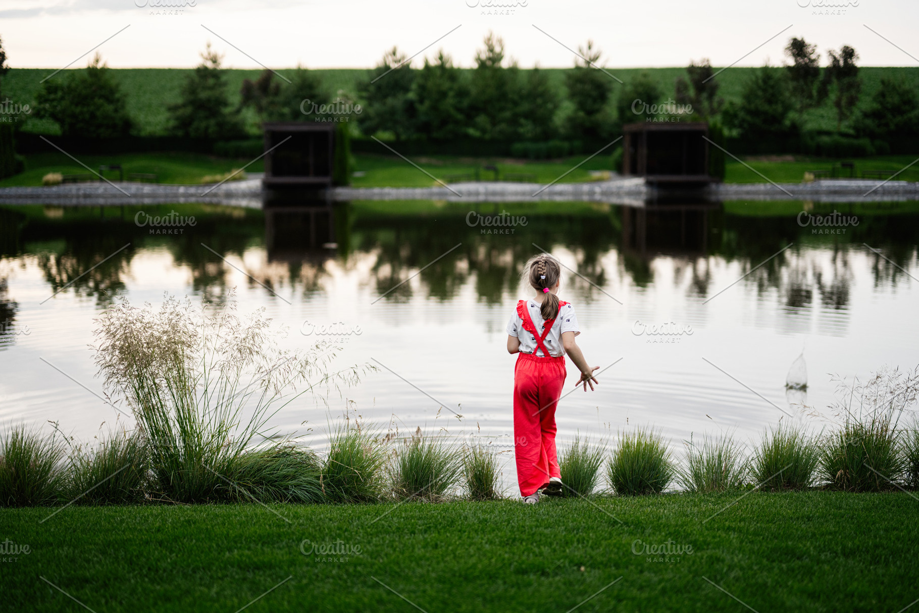 Young girl in red pants gazing at a serene pond in the evening, a Photo by AS photostudio